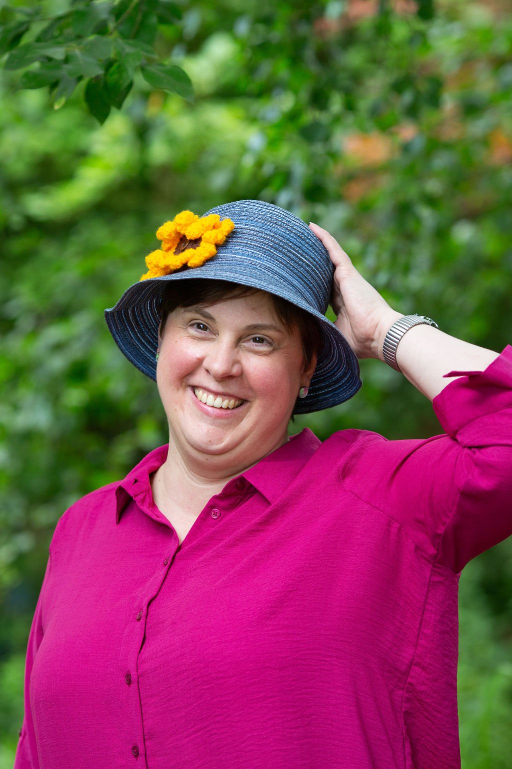 Lou, a white woman wearing a blue bucket hat with a knitted yellow flower attached. She is smiling at the camera and has one hand behind her head.