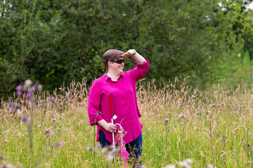 Lou, a white woman with short dark hair, wearing a pink top, outside in a field of purple flowers and long grass, holding her red and white cane and wearing dark glasses, looking into the distance with her hand held above her eyes to sheild her from the sun