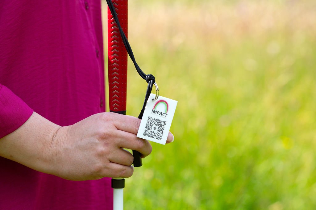 A zoomed in image of Lou's hand, holding her red and white cane with a keyring for Impact with Lou and a QR code.