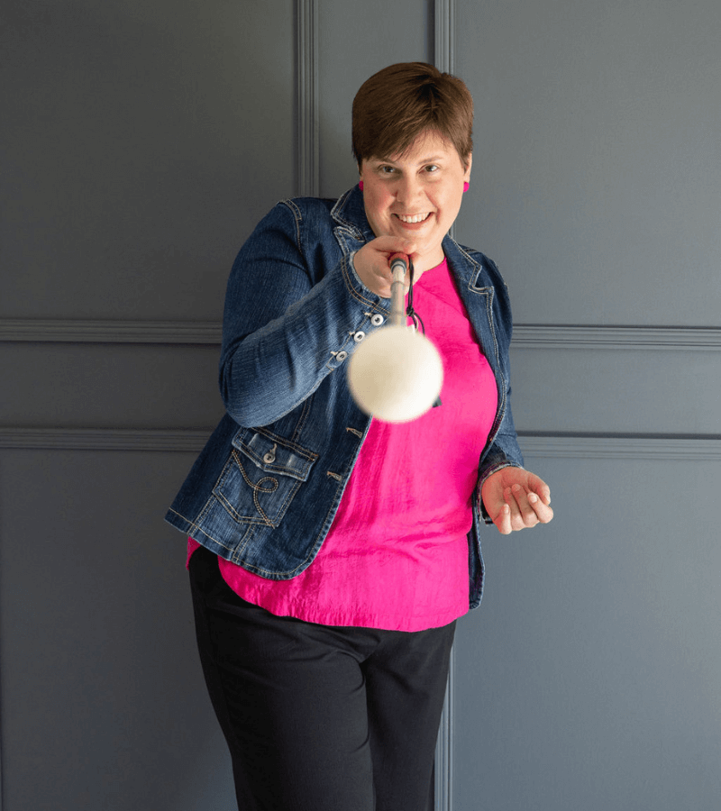 Lou, a white woman with short dark hair, wearing a pink top with a denim jacket. She is smiling and holding a cane towards the camera