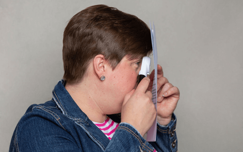 Lou, sat at a desk looking through a magnifying eyepiece to read something on a notepad close to her face